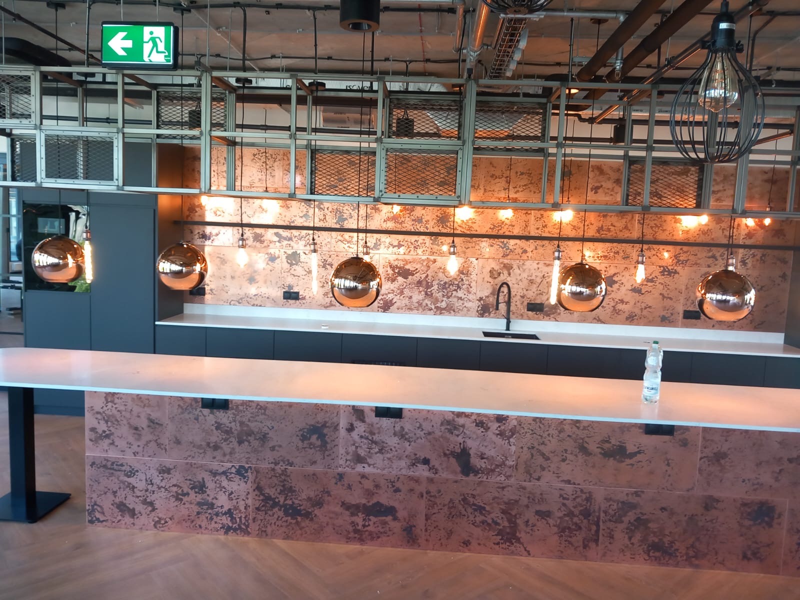 Nowoczesna kuchnia z blatem oświetlonym wiszącymi lampami. Miedziane wykończenia ścian i frontów szafek. Widoczny fragment podłogi z parkietu ułożonego w jodełkę.