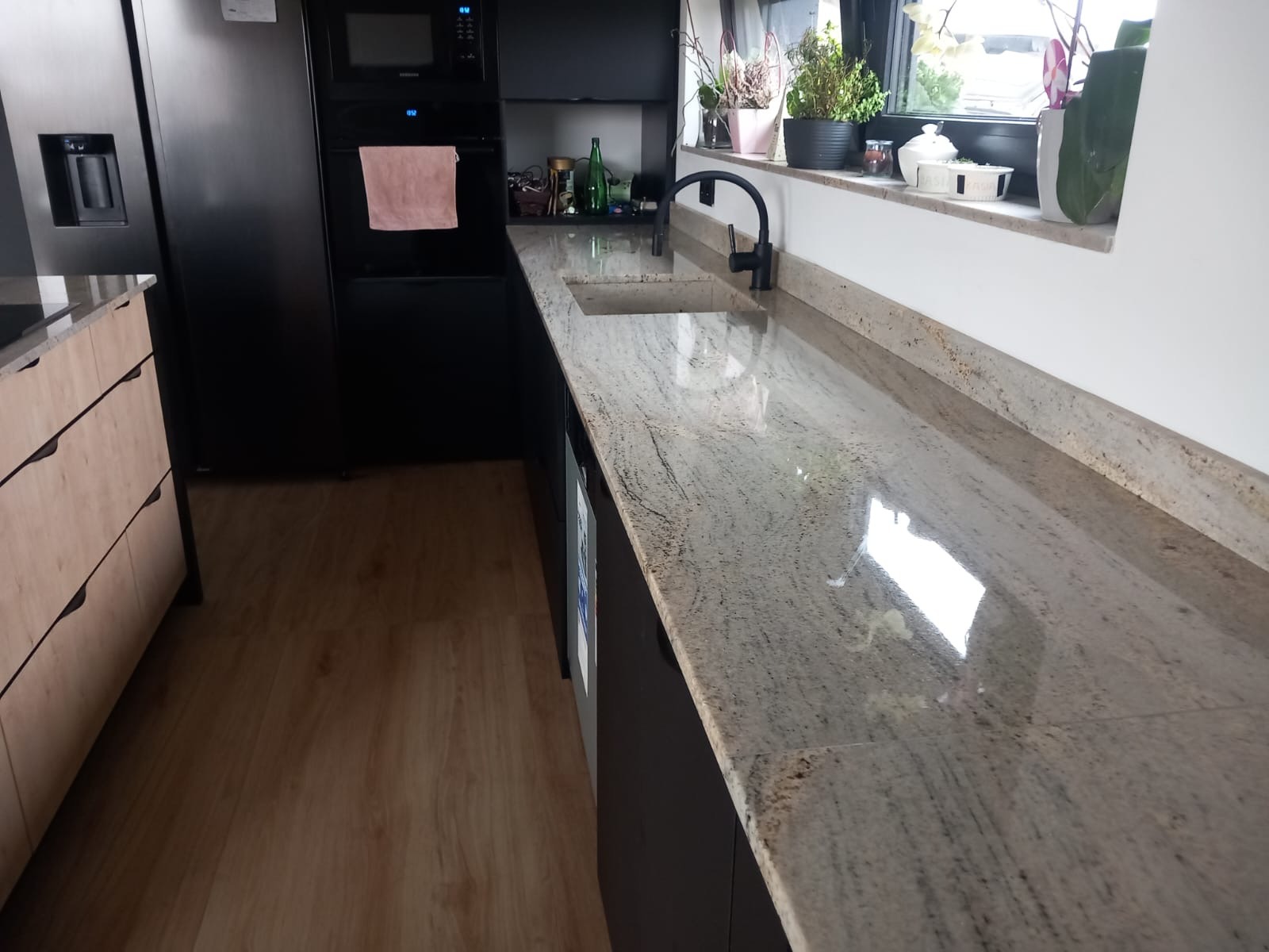 Długi, polerowany blat kamienny z wbudowanym zlewem w nowoczesnej kuchni z czarnymi szafkami i drewnianą podłogą. Widoczne rośliny w oknie.