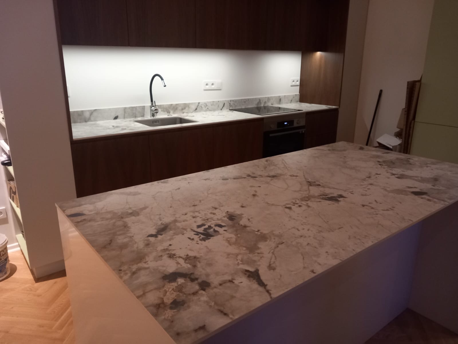 Kuchnia z wyspą i blatem z kamienia naturalnego o unikalnym wzorze żyłkowania. Ciemne szafki i jasne oświetlenie nad blatem roboczym.