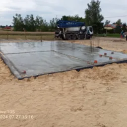 Zalany betonem fundament z wystającymi prętami zbrojeniowymi, widoczna czarna folia izolacyjna, w tle betoniarka i grupa osób.