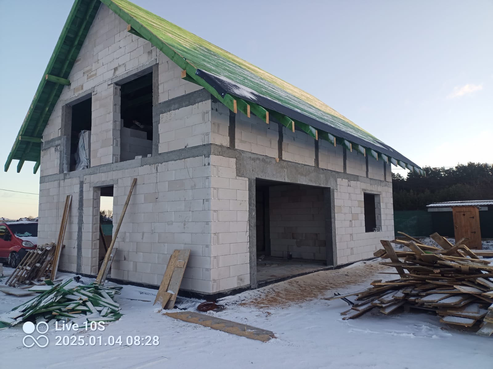 Niedokończony budynek z białych bloczków, z zielonym dachem w trakcie budowy, widoczne deski i śnieg na ziemi, zarys samochodu w tle.