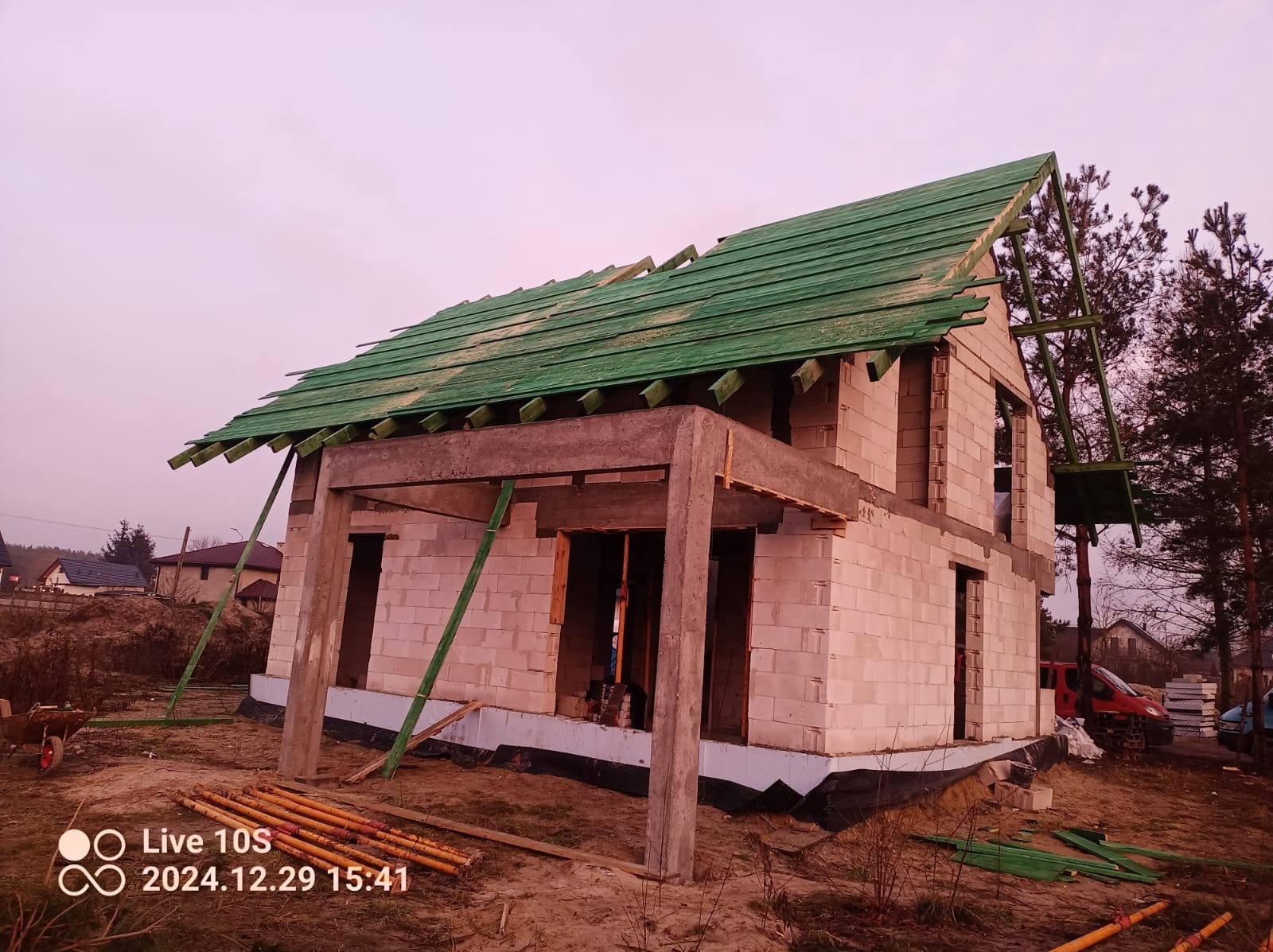 Surowy stan budynku z białego bloczka z zielonym pokryciem dachowym w trakcie budowy, widoczne niezabezpieczone otwory okienne i drzwiowe, teren budowy.