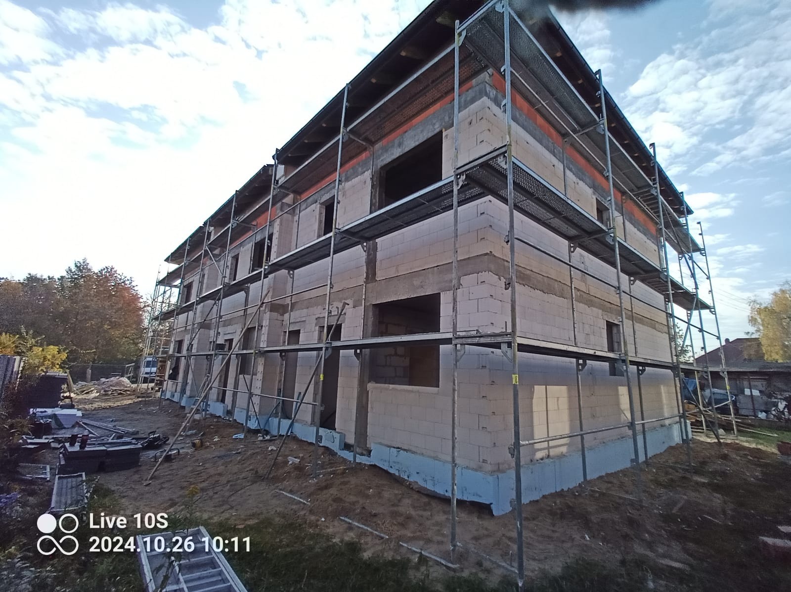 Trzypiętrowy budynek w trakcie budowy z białych bloczków, otoczony metalowym rusztowaniem, widoczne zabezpieczone fundamenty, niebo z chmurami w tle.