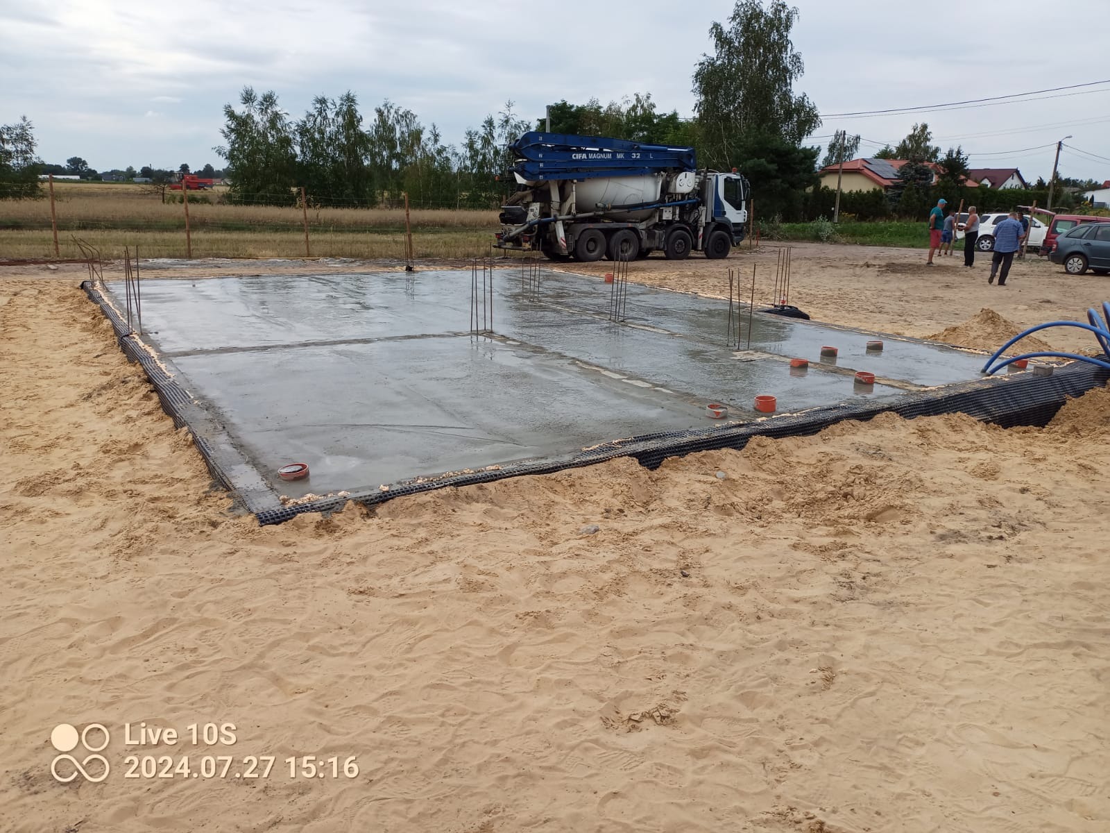 Zalany betonem fundament z wystającymi prętami zbrojeniowymi, widoczna czarna folia izolacyjna, w tle betoniarka i grupa osób.