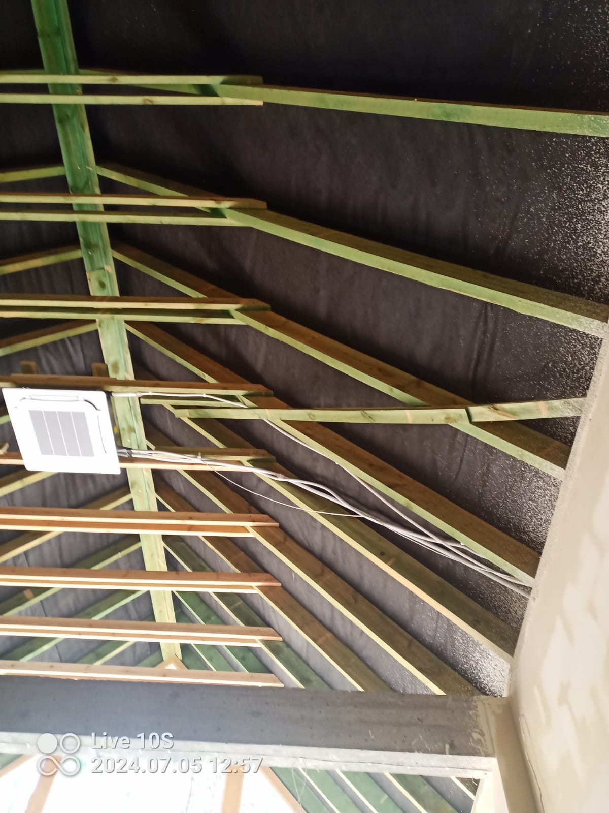 Konstrukcja dachu z widoczną membraną dachową, drewnianymi belkami pomalowanymi na zielono, przewodami elektrycznymi i zamontowaną klimatyzacją, widok z dołu.