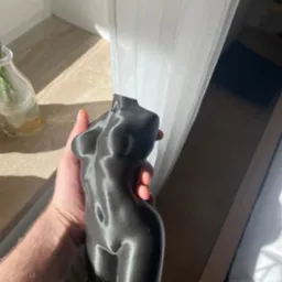 Czarna, połyskująca figurka kobiecego torsu, wydrukowana w 3D, trzymana w dłoni na tle okna z firanką i blatem. Widoczne refleksy światła.