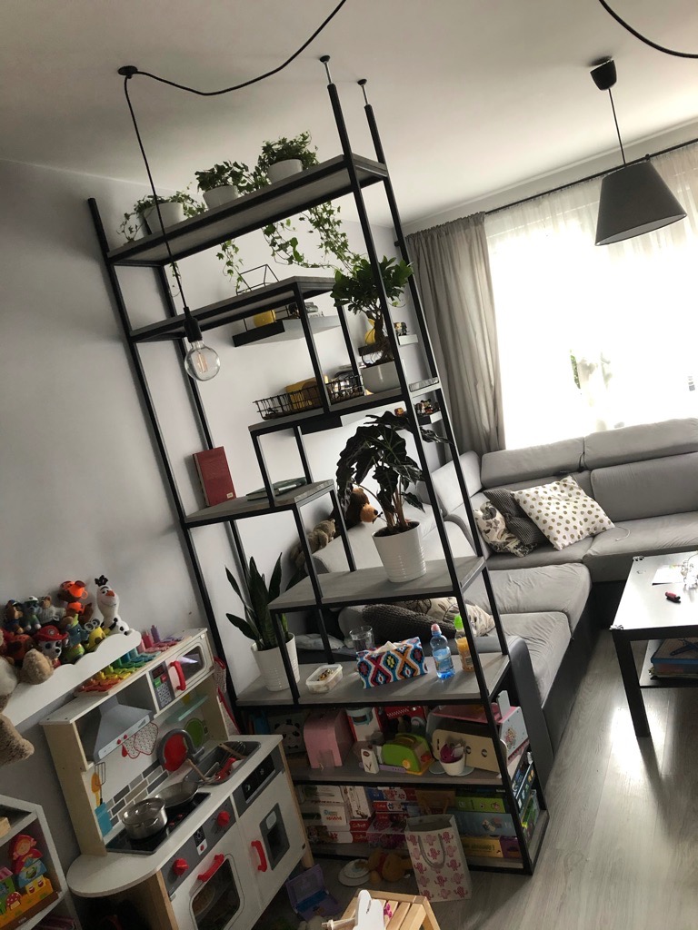 Metalowy regał z półkami, wykorzystany jako ścianka działowa w salonie, z roślinami i dekoracjami. Widoczna dziecięca kuchnia i szara sofa w tle.