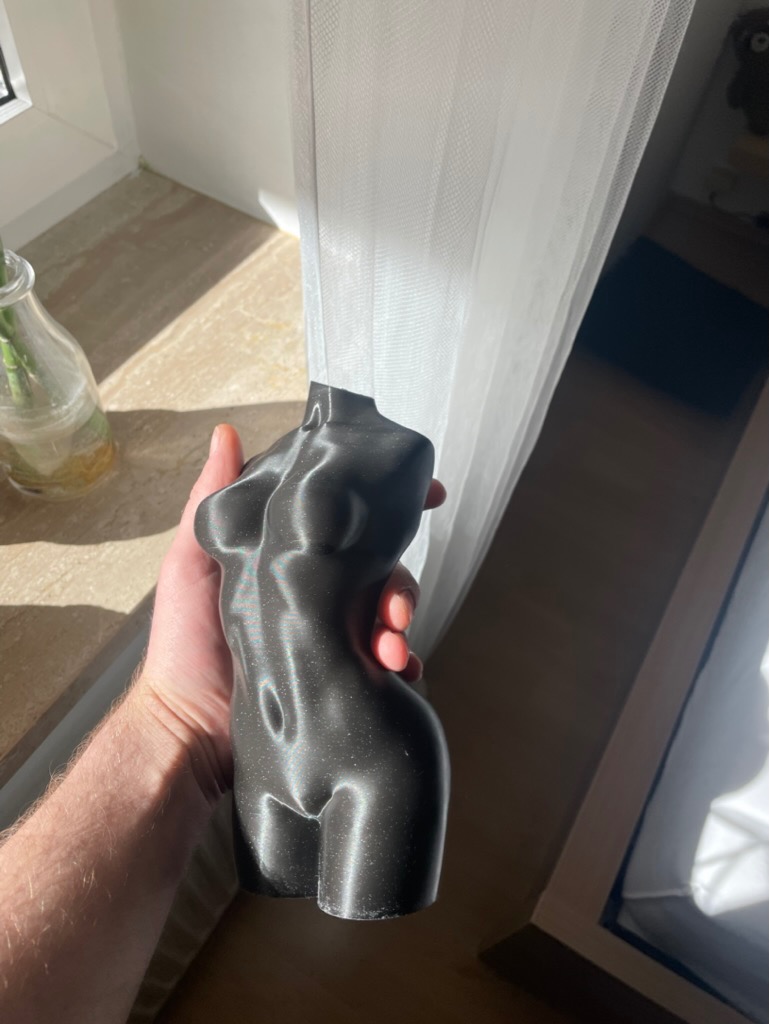Czarna, połyskująca figurka kobiecego torsu, wydrukowana w 3D, trzymana w dłoni na tle okna z firanką i blatem. Widoczne refleksy światła.