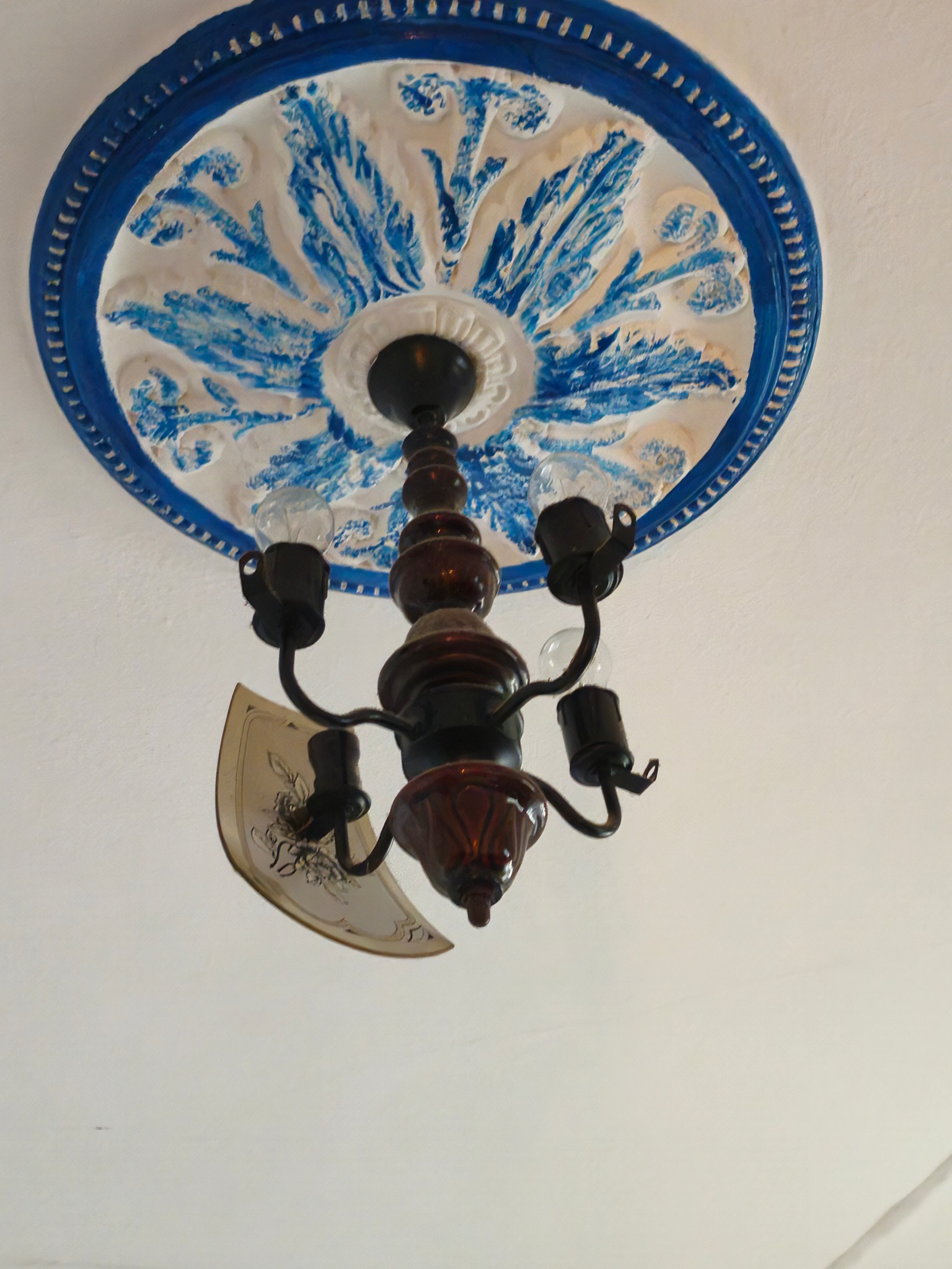 Dekoracyjny rozetowy sufit z niebieskimi akcentami malarskimi, z którego zwisa zabytkowy żyrandol z czterema żarówkami i ozdobnymi kloszami.