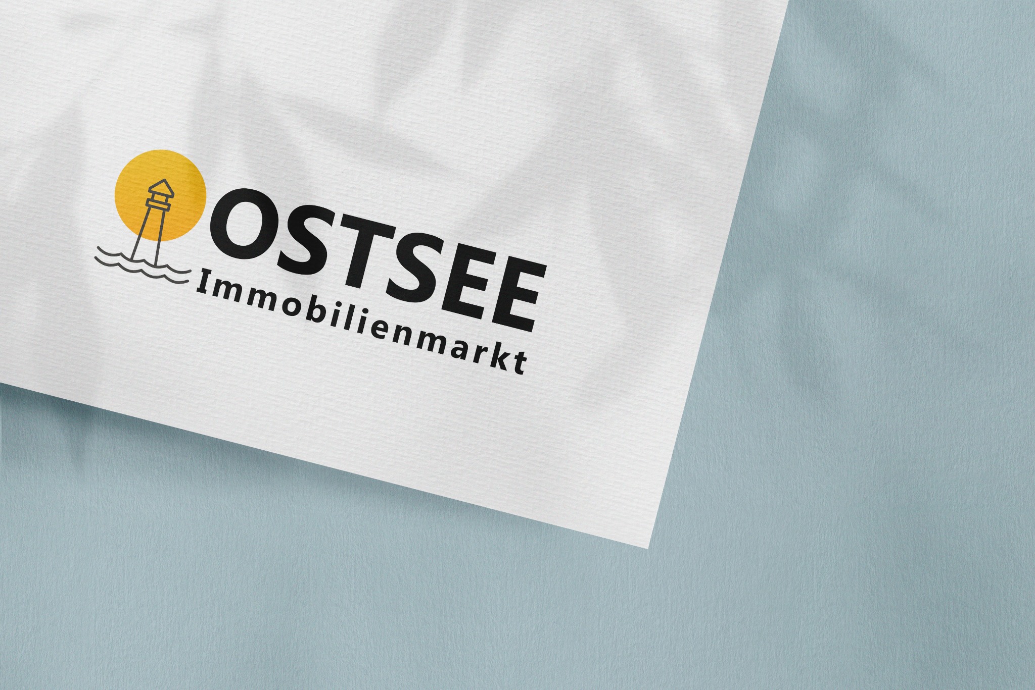 Graficzne logo firmy Ostsee Immobilienmarkt przedstawiające latarnię morską na tle słońca, umieszczone na białym papierze z cieniem liści na niebieskim tle.
