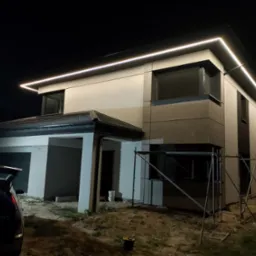 Nowoczesny dom jednorodzinny z oświetleniem LED elewacji, widoczne rusztowania i teren w trakcie prac wykończeniowych, ciemne tło nocne.