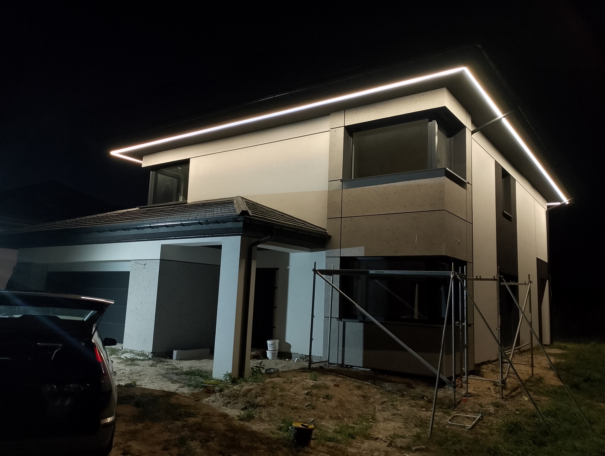 Nowoczesny dom jednorodzinny z oświetleniem LED elewacji, widoczne rusztowania i teren w trakcie prac wykończeniowych, ciemne tło nocne.