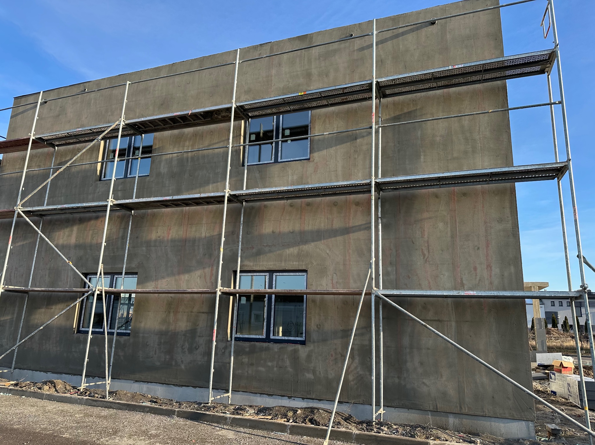 Budynek w trakcie ocieplania, widoczne rusztowania i surowa elewacja z oknami, przygotowana pod dalsze prace wykończeniowe.