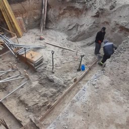 Redar - Wykop pod budowę domu jednorodzinnego: dwóch pracowników w wykopie, szpadle, poziomica, przygotowane szalunki, hałda ziemi w tle.