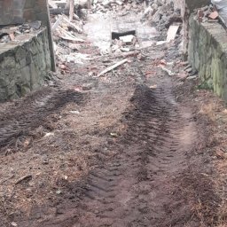 Redar - Teren po wyburzeniu budynku z widocznymi śladami opon ciężkiego sprzętu, gruz ceglany i fragmenty ścian, w tle las i obserwujący robotników mężczyźni.