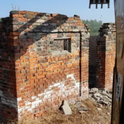 Resztki muru z czerwonej cegły z widocznym fragmentem koparki, otoczone gruzem i suchą trawą, na tle błękitnego nieba.