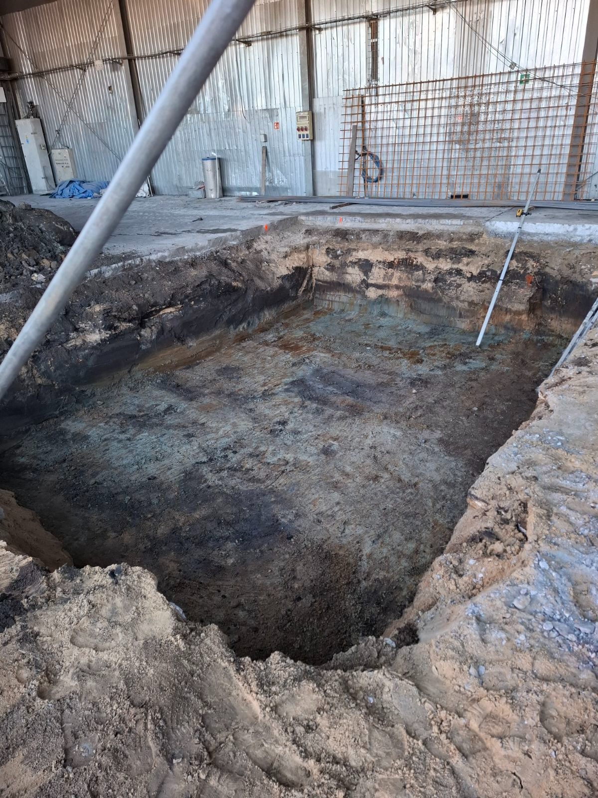 Głęboki wykop w ziemi wewnątrz hali, widoczne warstwy gruntu i poziomica, przygotowany pod budowę fundamentów.