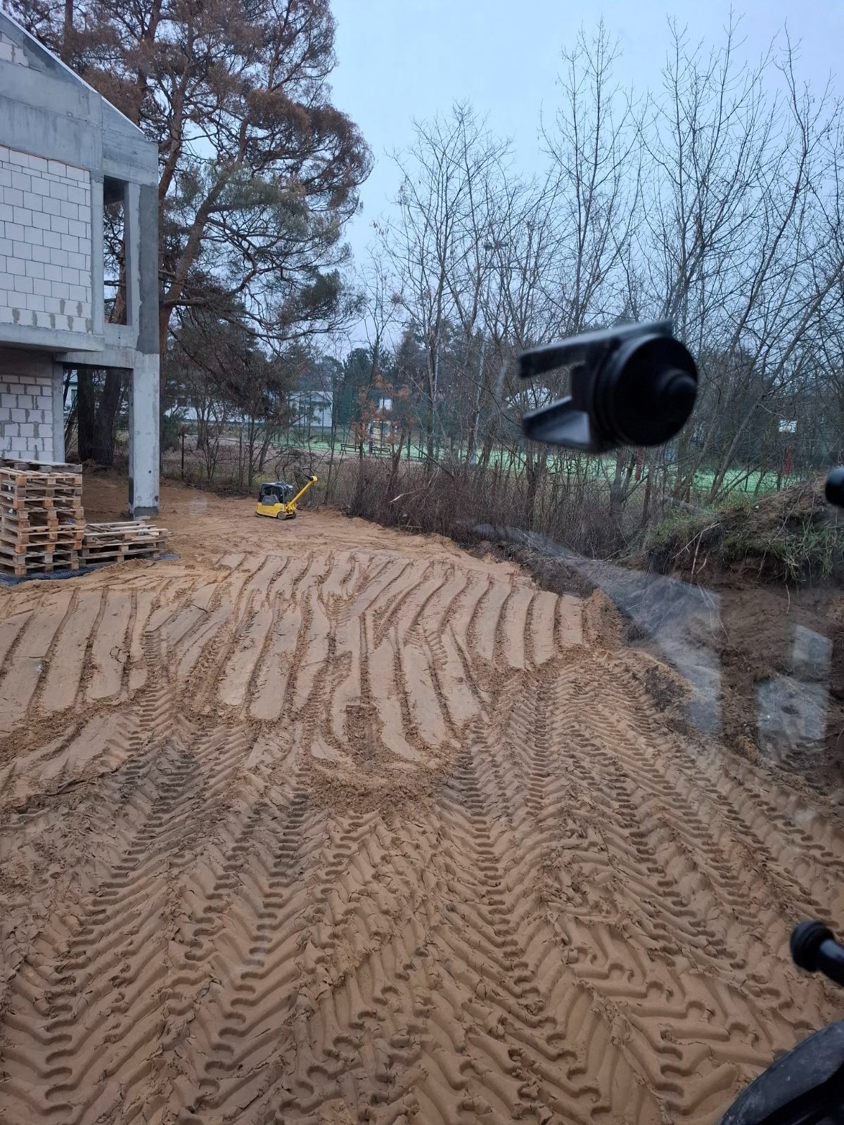 Przygotowany teren pod budowę domu jednorodzinnego z widocznymi śladami po zagęszczarce, palety z bloczkami fundamentowymi i mała koparka w tle.