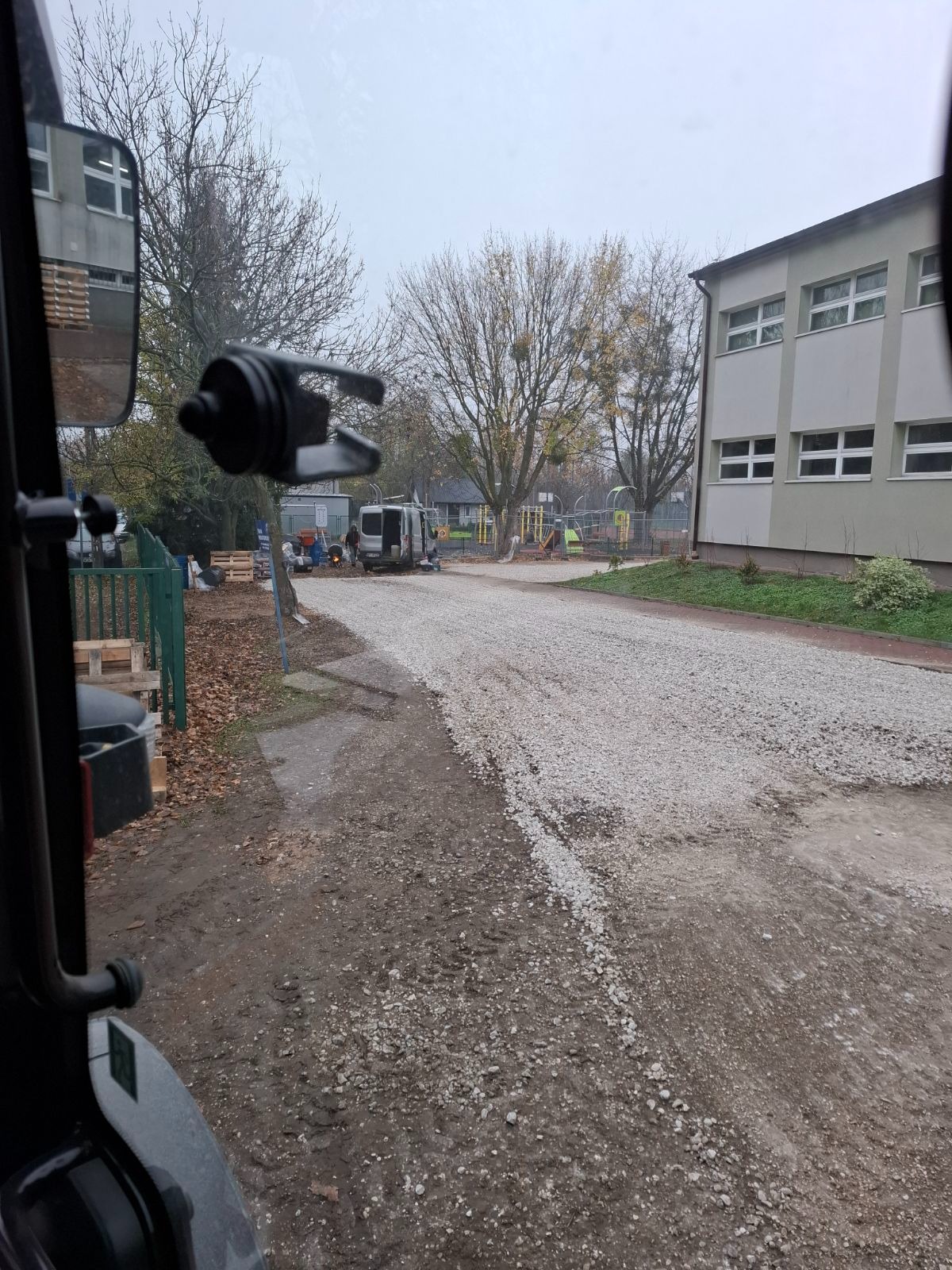Widok z kabiny maszyny budowlanej na teren przygotowywany pod drogę, z rozłożonym kruszywem; w tle budynek, drzewa i ekipa robotników przy furgonetce.