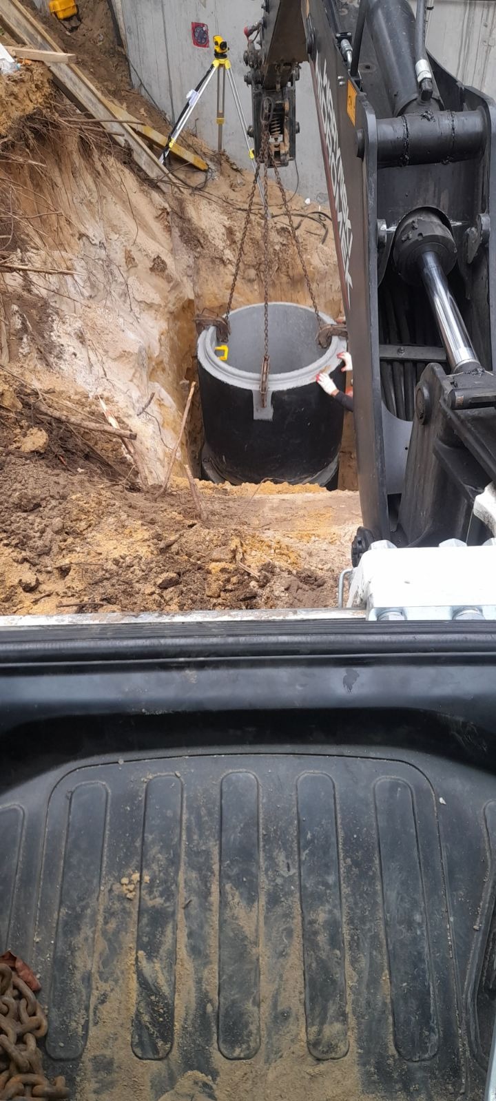 Ciężki sprzęt opuszcza betonowy zbiornik do wykopu w ziemi, widoczny niwelator laserowy na statywie.