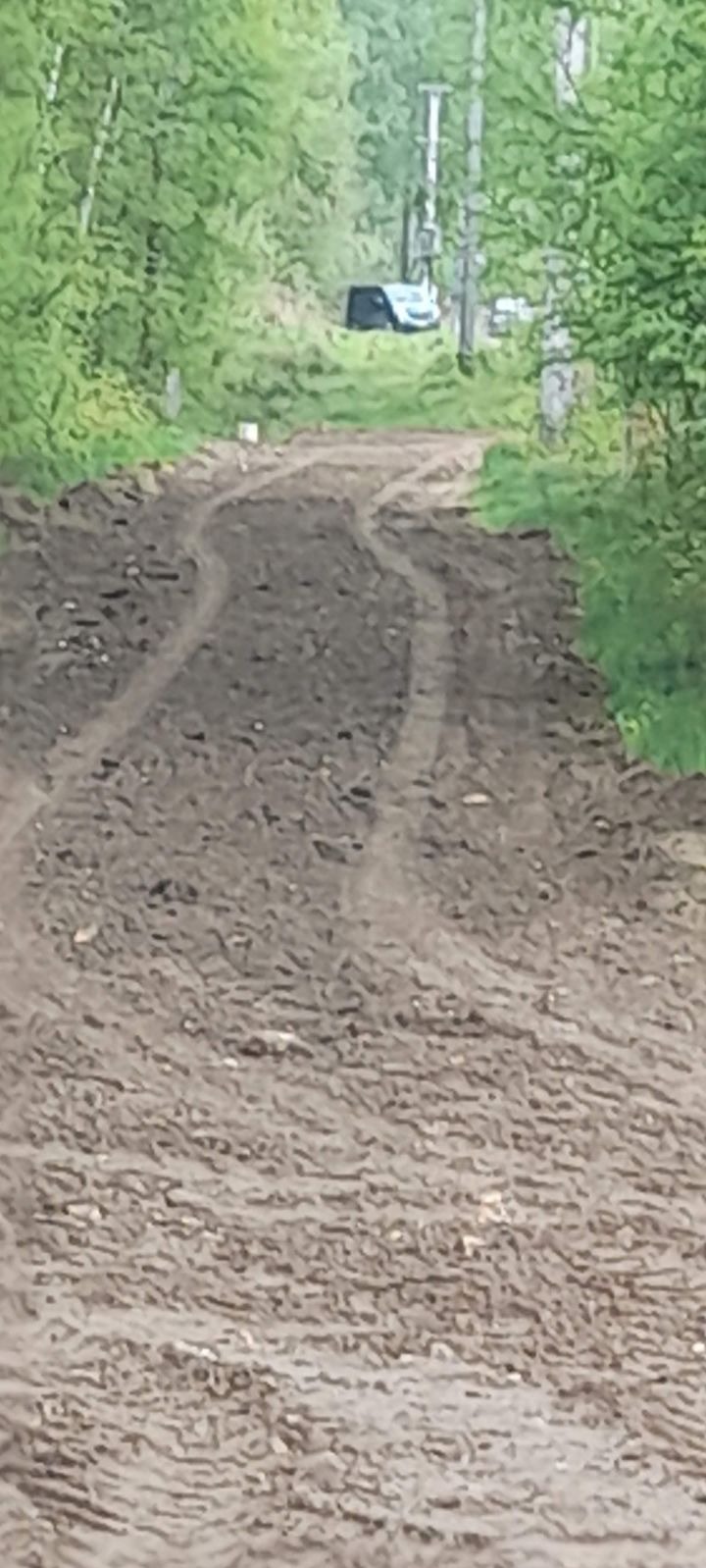 Świeżo wyrównana, błotnista droga gruntowa z widocznymi śladami opon w lesie, w tle słup energetyczny i budynek.