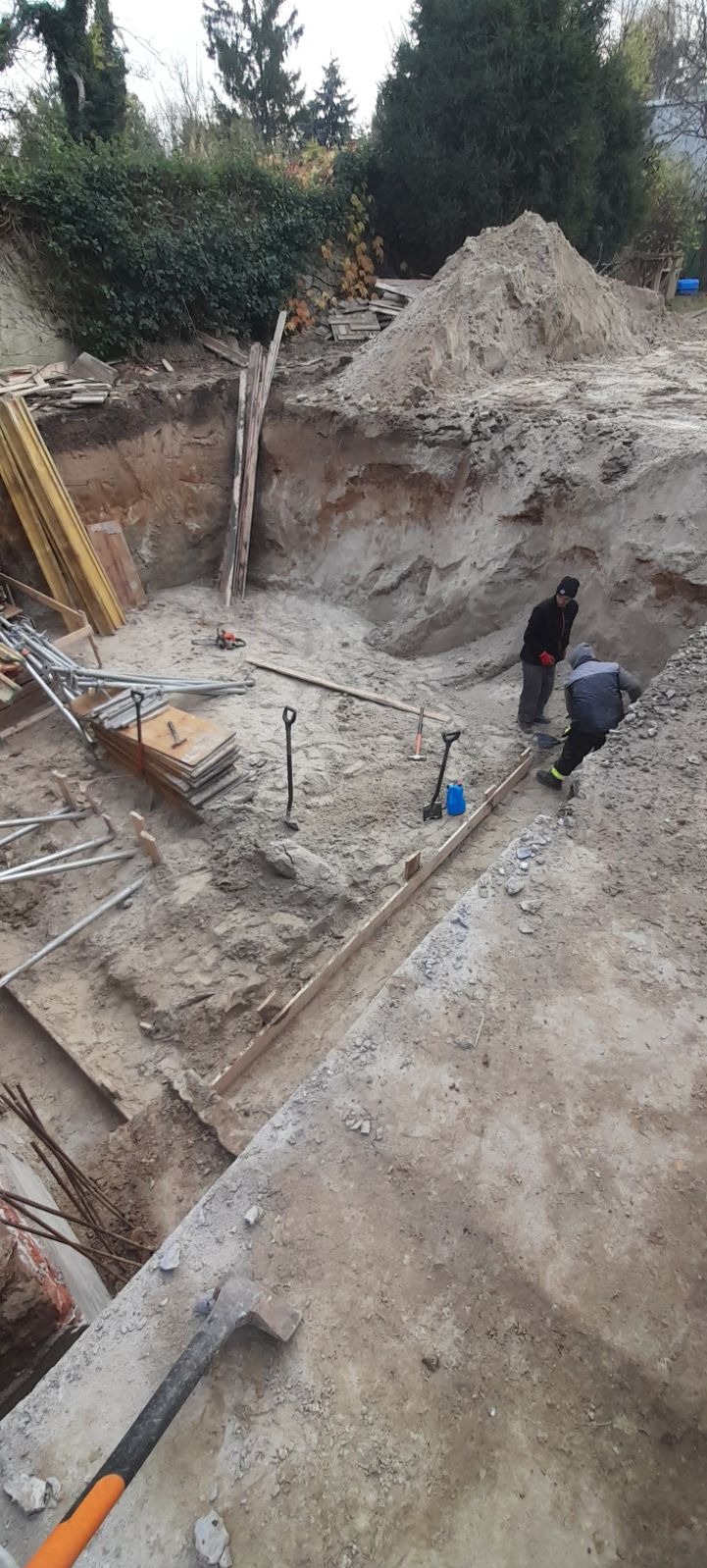 Wykop pod budowę domu jednorodzinnego: dwóch pracowników w wykopie, szpadle, poziomica, przygotowane szalunki, hałda ziemi w tle.