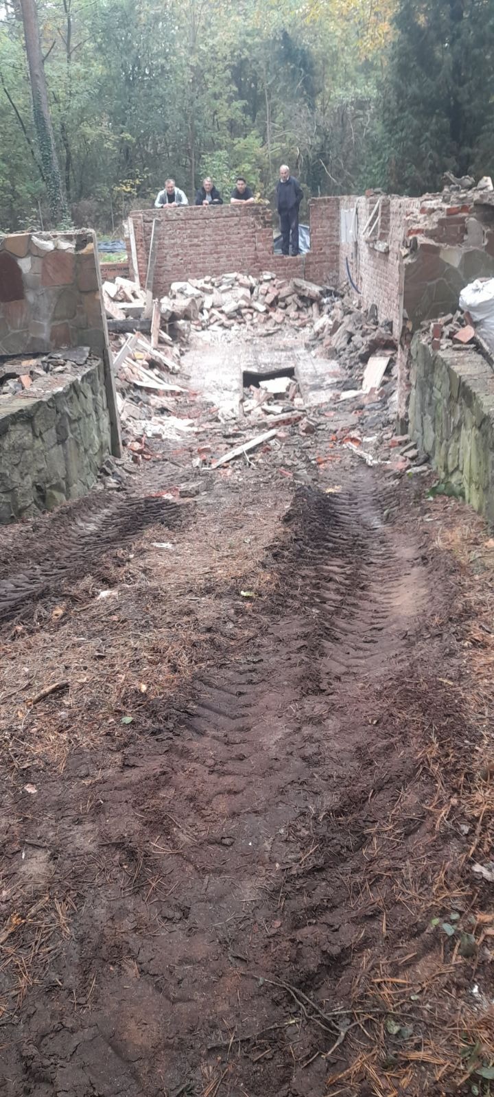 Teren po wyburzeniu budynku z widocznymi śladami opon ciężkiego sprzętu, gruz ceglany i fragmenty ścian, w tle las i obserwujący robotników mężczyźni.