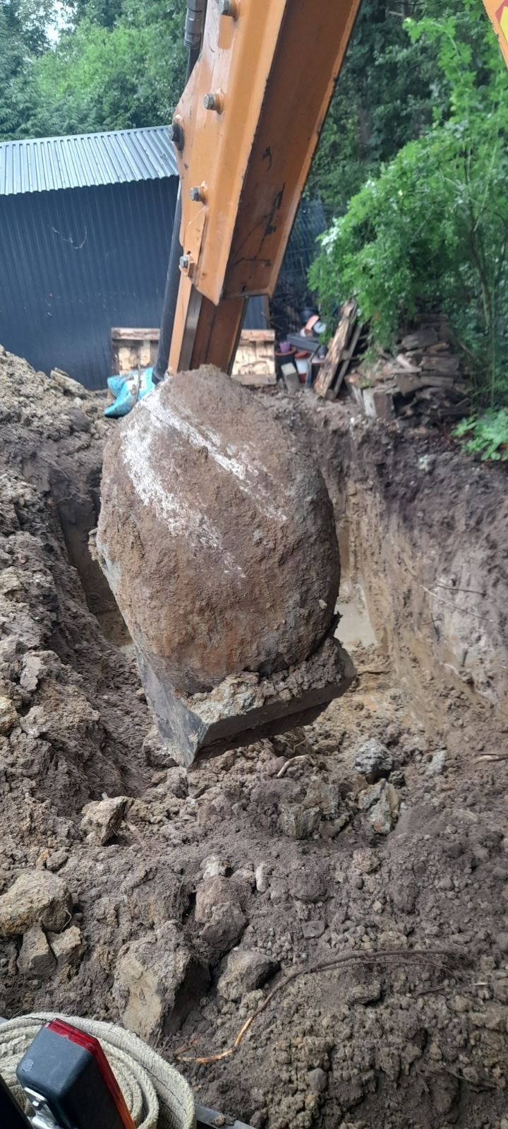 Pomarańczowa koparka z łyżką podnosi duży, okrągły kamień z wykopu w ziemi, w tle widoczny budynek z blachy falistej i zieleń.