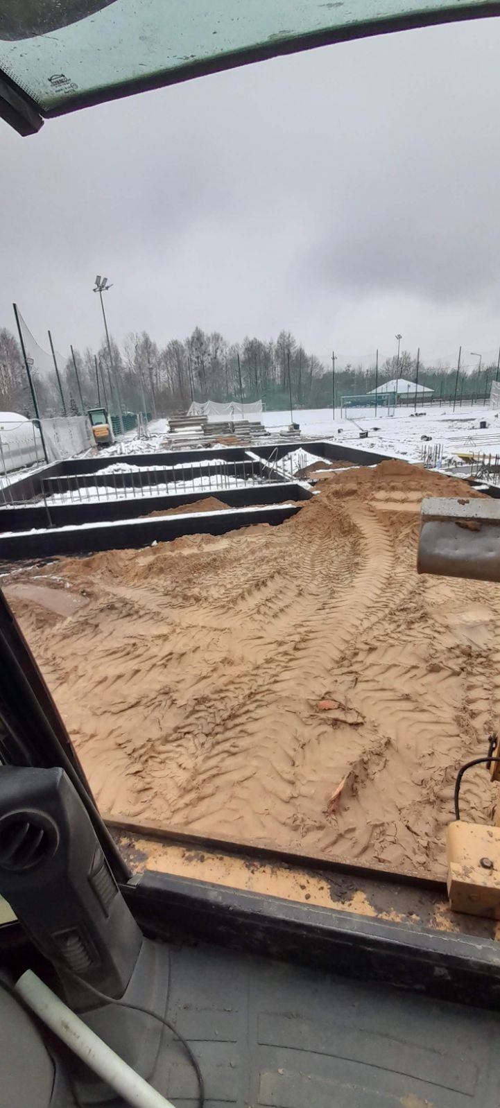 Widok z kabiny maszyny budowlanej na plac budowy w zimowej scenerii; widoczne wykopane fundamenty zasypane piaskiem, ślady opon, w tle częściowo ośnieżone boisko sportowe.