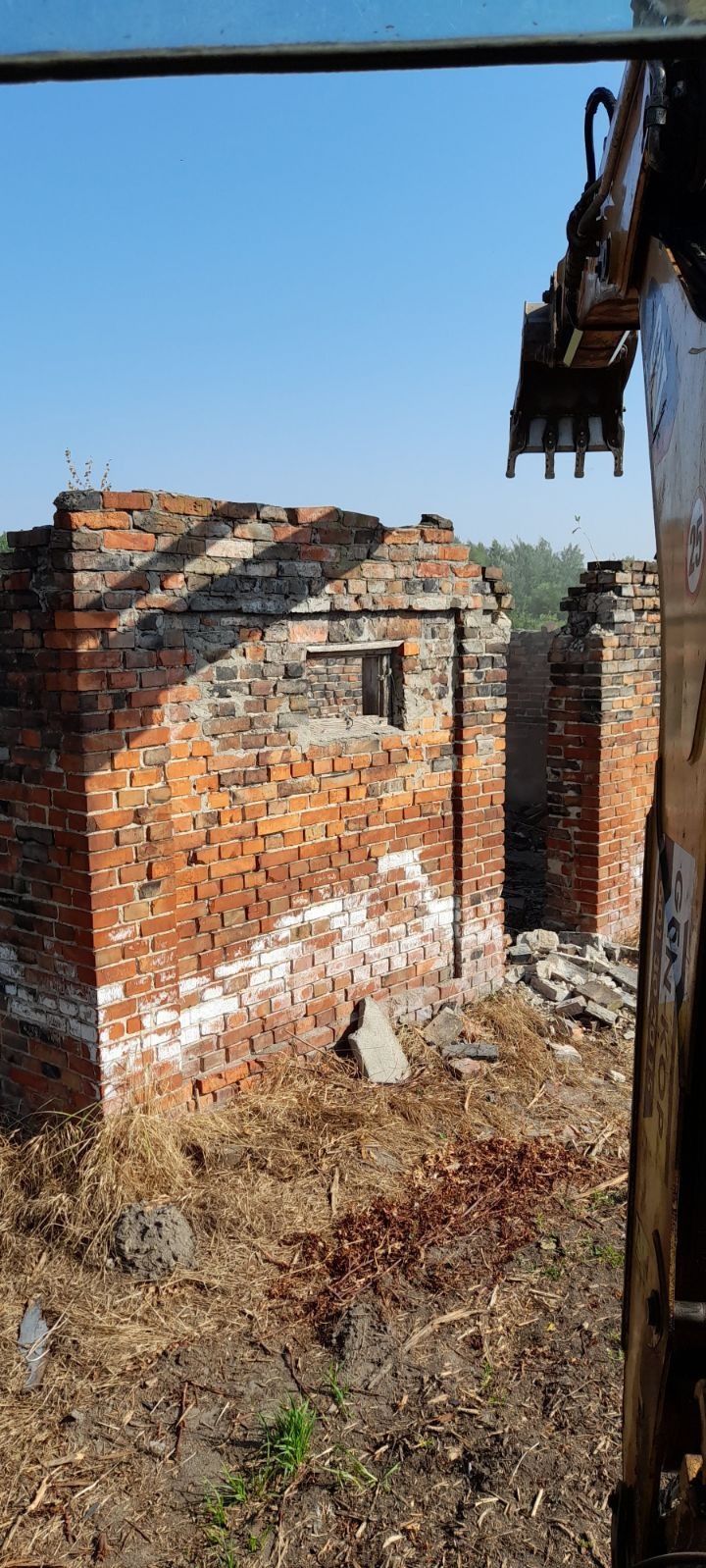 Resztki muru z czerwonej cegły z widocznym fragmentem koparki, otoczone gruzem i suchą trawą, na tle błękitnego nieba.