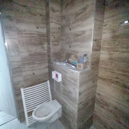 Krzysztof Balcerzak - Wykończona łazienka z beżowymi płytkami imitującymi drewno, wiszącym bidetem i grzejnikiem drabinkowym. Widoczne materiały budowlane na półce.