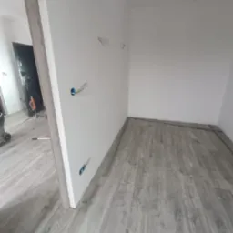 Wnętrze w trakcie układania paneli podłogowych w odcieniach szarości. Widoczne kable wystające ze ściany i fragmenty listew przypodłogowych.