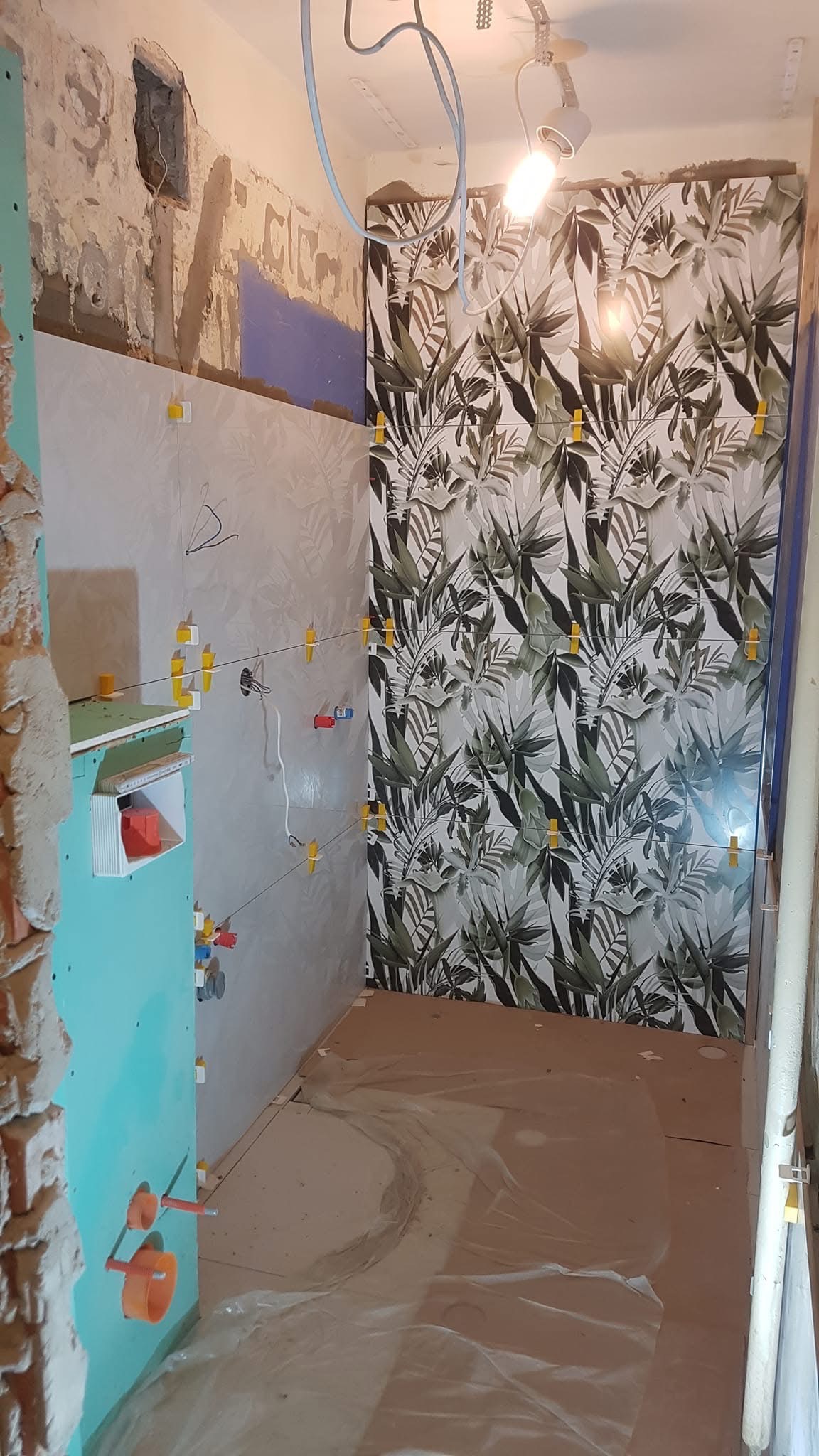 Układanie płytek z motywem roślinnym w łazience, widoczne krzyżyki dystansowe, fragment instalacji wodnej i niedokończone ściany. Prace wykończeniowe w toku.