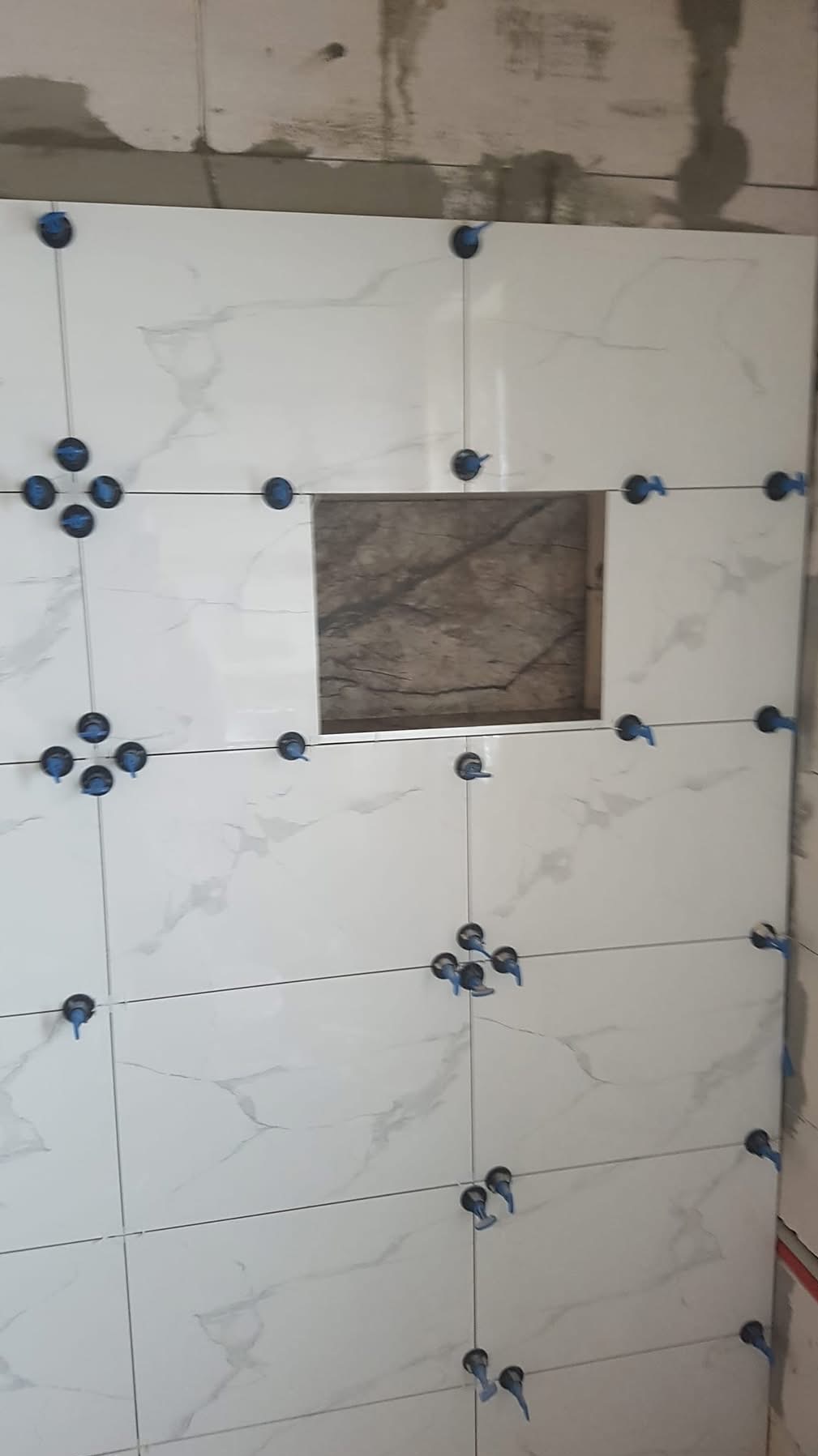 Ściana z białych płytek imitujących marmur, z wnęką wyłożoną ciemniejszymi płytkami. Użyto niebieskich krzyżyków dystansowych do fugowania.