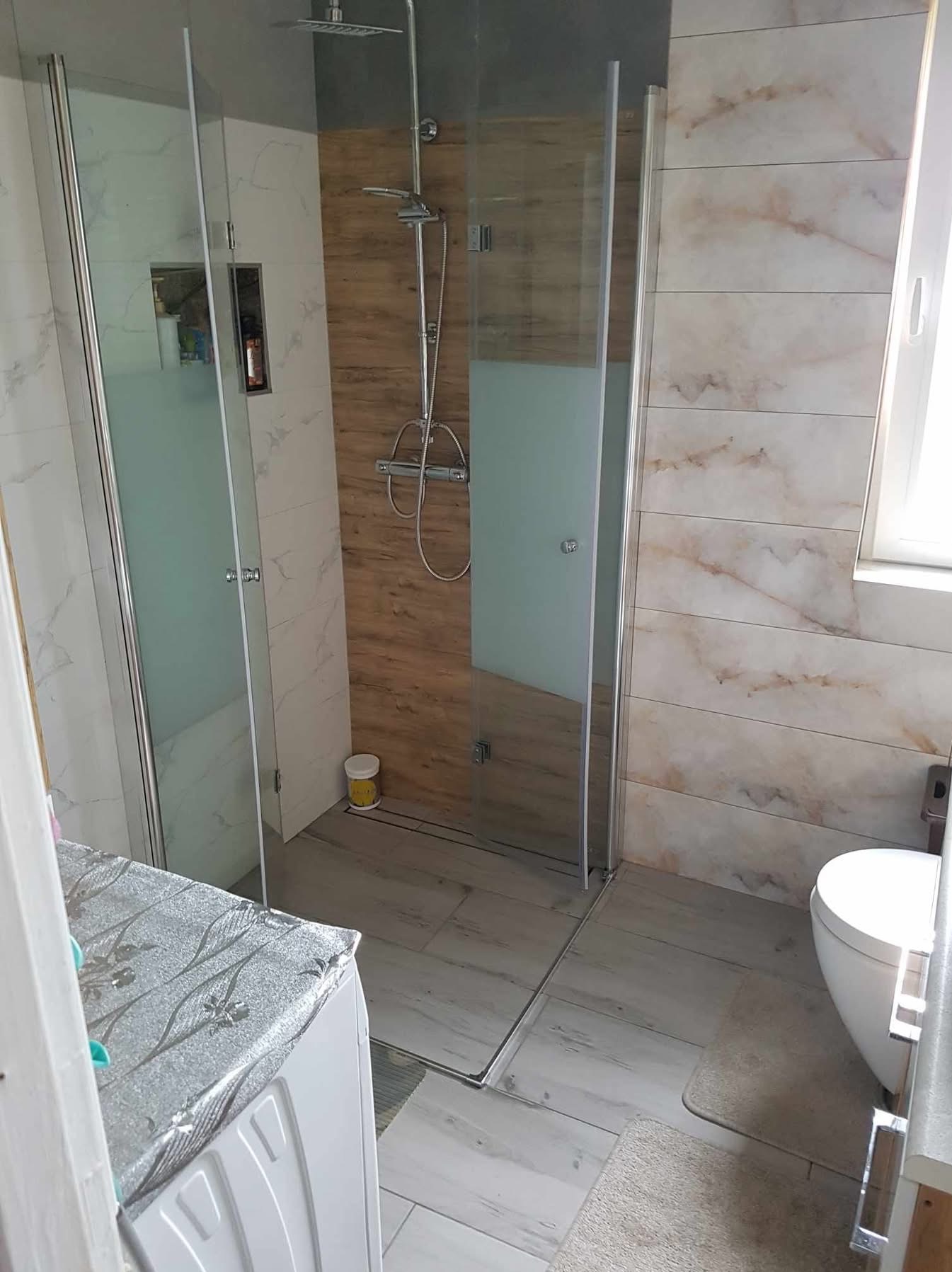 Nowoczesna kabina prysznicowa z deszczownicą i szklanymi drzwiami, wykończona płytkami imitującymi drewno i marmur. Widoczna toaleta i fragment pralki.