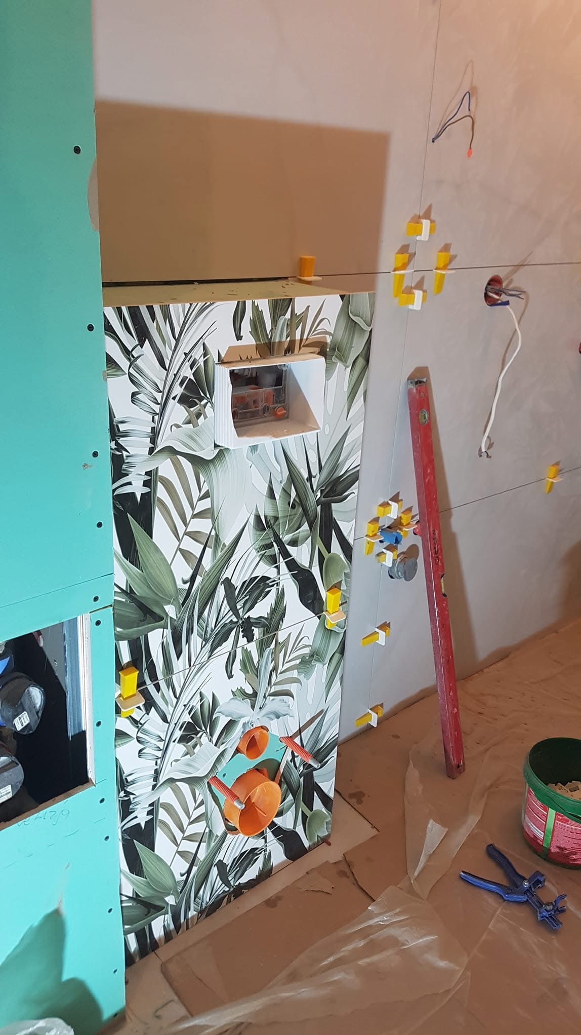 Montaż płytek z motywem roślinnym w łazience, widoczne narzędzia i poziomica. Częściowo zamontowana płyta karton gips. Etap prac wykończeniowych.