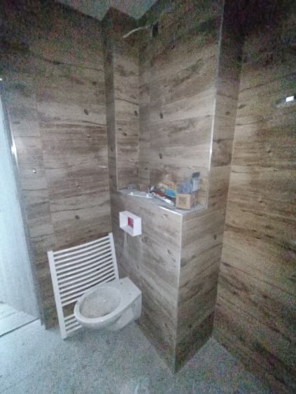 Wykończona łazienka z beżowymi płytkami imitującymi drewno, wiszącym bidetem i grzejnikiem drabinkowym. Widoczne materiały budowlane na półce.