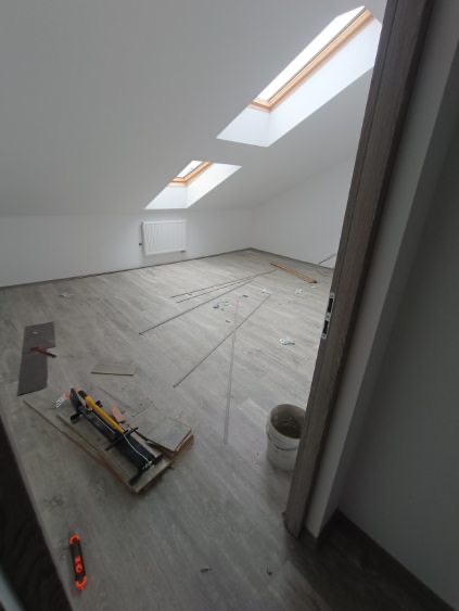 Układanie paneli podłogowych w jasnym pokoju na poddaszu ze skosami i oknami dachowymi. Widoczny fragment grzejnika i narzędzia do cięcia paneli.