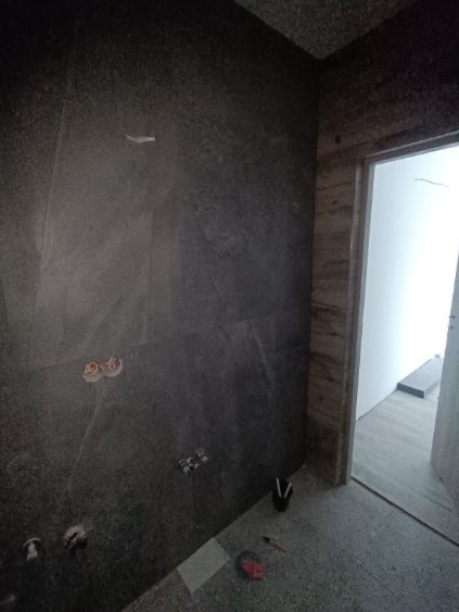 Wykończona ściana z ciemnej, strukturalnej płyty w łazience. Widoczne przyłącza wodne i fragment podłogi z narzędziami. W tle ościeżnica drzwi.