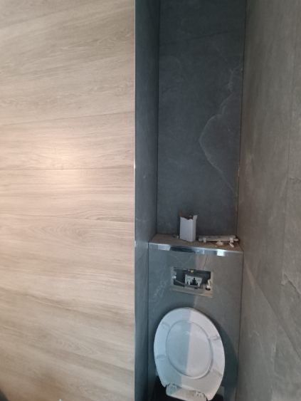 Nowoczesna toaleta podwieszana w zabudowie z szarych płytek, z widoczną instalacją i elementami montażowymi. Ściana wykończona płytkami imitującymi drewno.