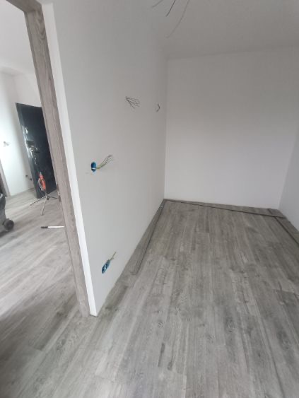 Wnętrze w trakcie układania paneli podłogowych w odcieniach szarości. Widoczne kable wystające ze ściany i fragmenty listew przypodłogowych.