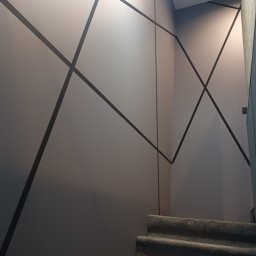 Damian Halabowski - Ściana klatki schodowej z geometrycznym wzorem z ciemnych pasów, szare schody pokryte wykładziną dywanową.