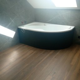 Damian Halabowski - Narożna wanna z czarną mozaiką na obudowie, szare płytki imitujące marmur na ścianach i podłoga wyłożona panelami imitującymi drewno w łazience ze skośnym sufitem i oknem dachowym.