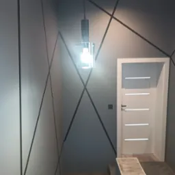 Nowoczesne wykończenie wnętrza: szare ściany z geometrycznym wzorem z czarnych linii, białe drzwi z poziomymi przeszkleniami i wisząca lampa z przezroczystym kloszem.