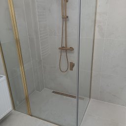 Damian Halabowski - Nowoczesna kabina prysznicowa z szklanymi drzwiami i złotymi profilami, szare płytki na ścianach i podłodze, odpływ liniowy w podłodze.