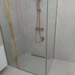 Nowoczesna kabina prysznicowa z szklanymi drzwiami i złotymi profilami, szare płytki na ścianach i podłodze, odpływ liniowy w podłodze.