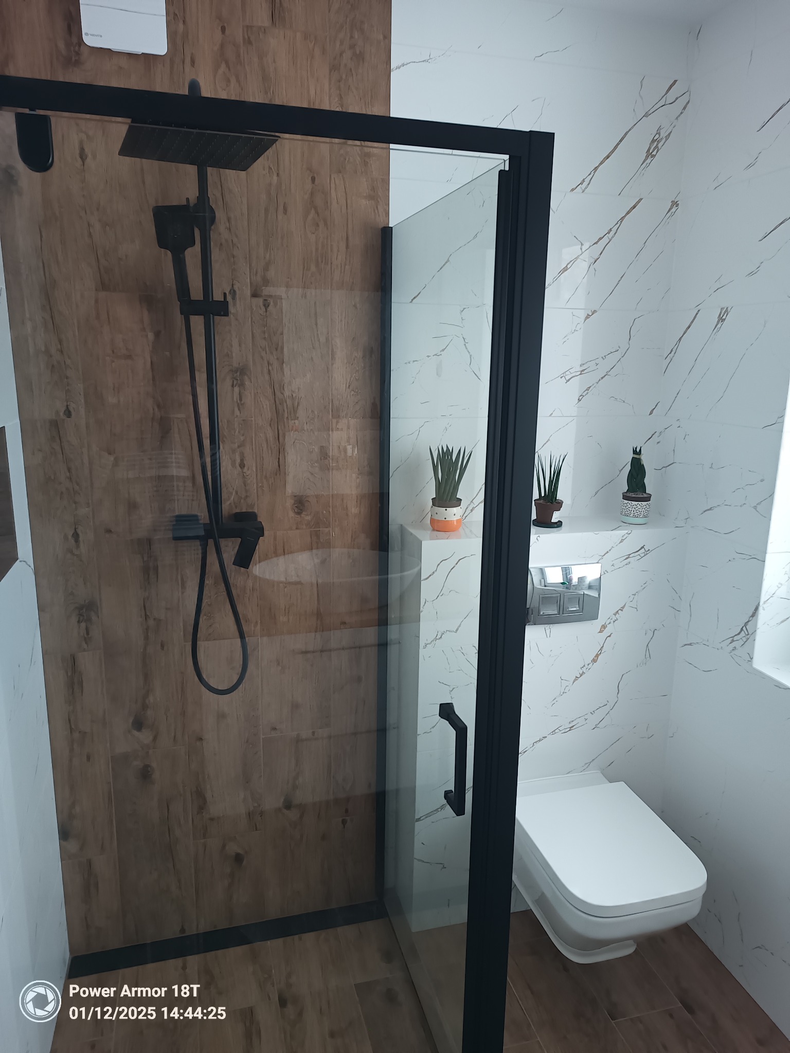 Nowoczesna kabina prysznicowa z czarną armaturą i drewnopodobnymi płytkami w strefie natrysku, wisząca toaleta z marmurowymi płytkami na ścianie i dekoracyjnymi roślinami doniczkowymi na półce.