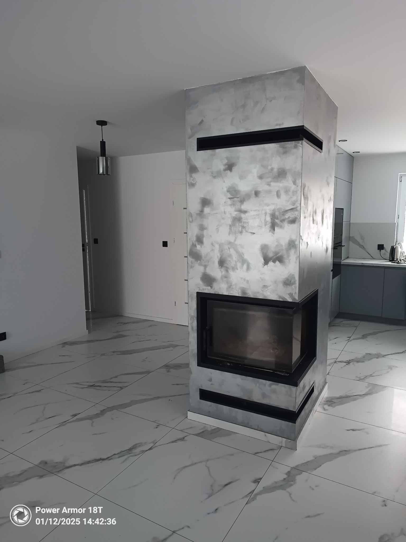 Nowoczesny kominek obudowany płytami imitującymi beton, z czarną ramą, w jasnym wnętrzu z marmurową podłogą i widoczną kuchnią w tle.