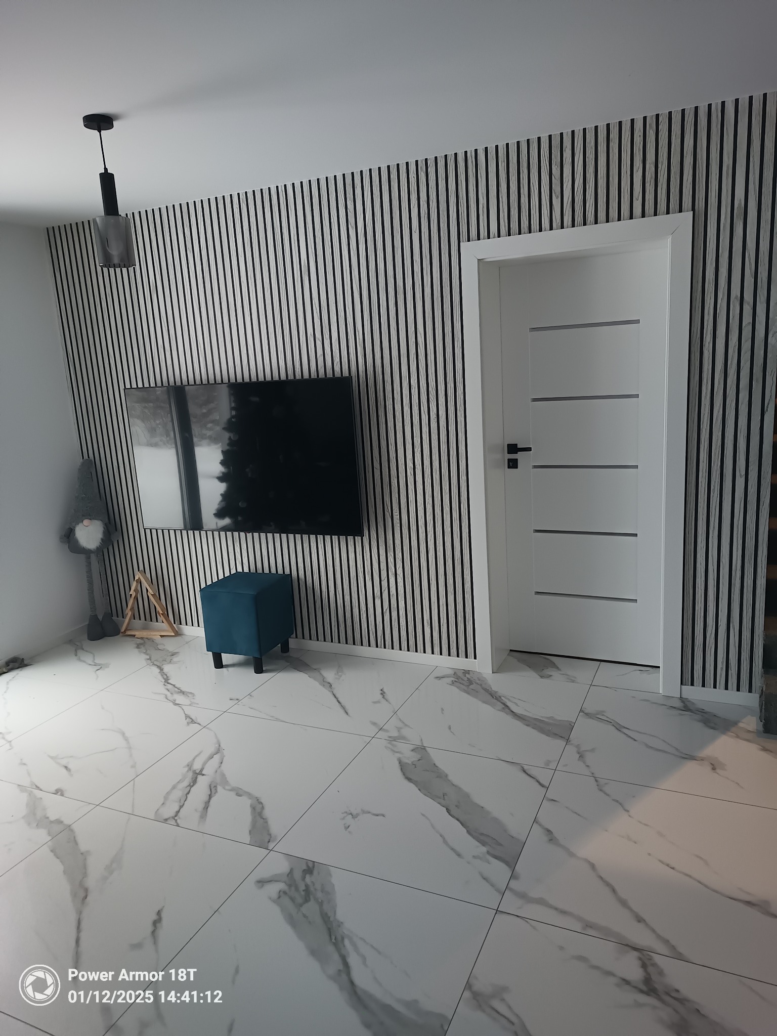 Wnętrze pokoju z białą, nowoczesną stolarką drzwiową, ścianą wykończoną panelami dekoracyjnymi imitującymi drewno, marmurową posadzką, niebieskim pufem i dekoracjami.