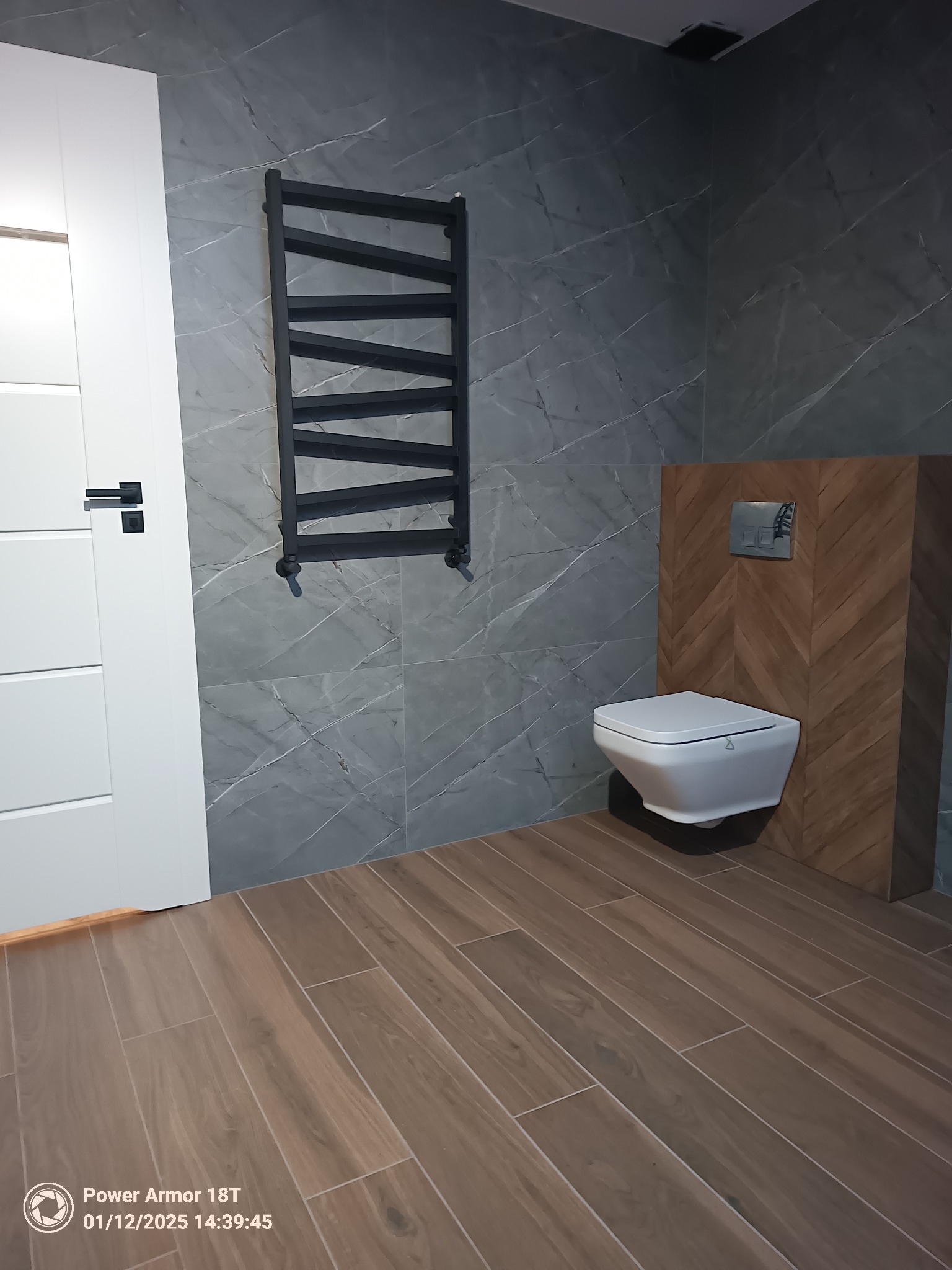 Nowoczesna łazienka z szarymi marmurkowymi płytkami ściennymi, drewnopodobną podłogą, wiszącą toaletą na tle drewnianej mozaiki i czarnym grzejnikiem drabinkowym.